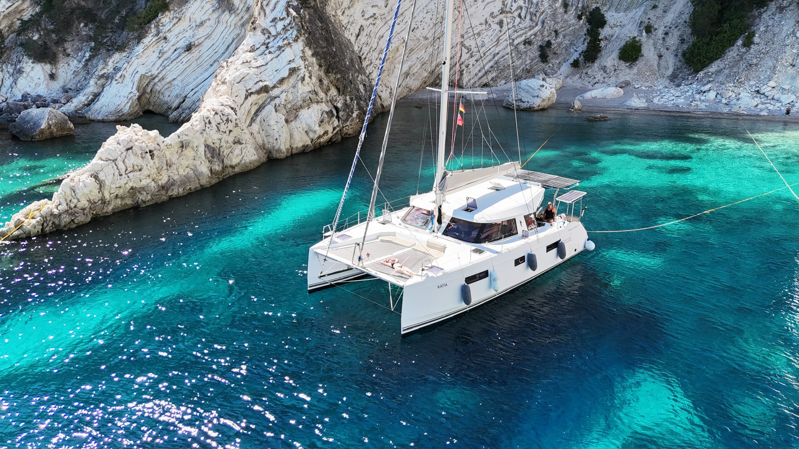 sailing catamaran katja in front of beach and cliff with bikini lady on foredeck