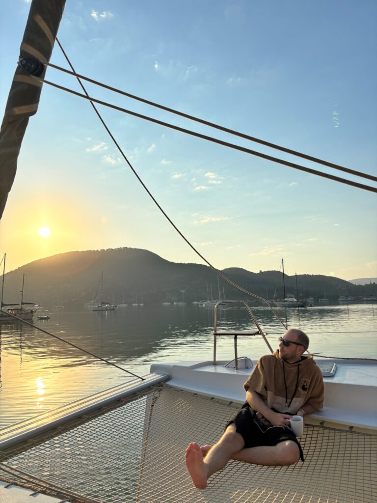 man with coffee mug sitting on catamran nets with sun dawn over island in background